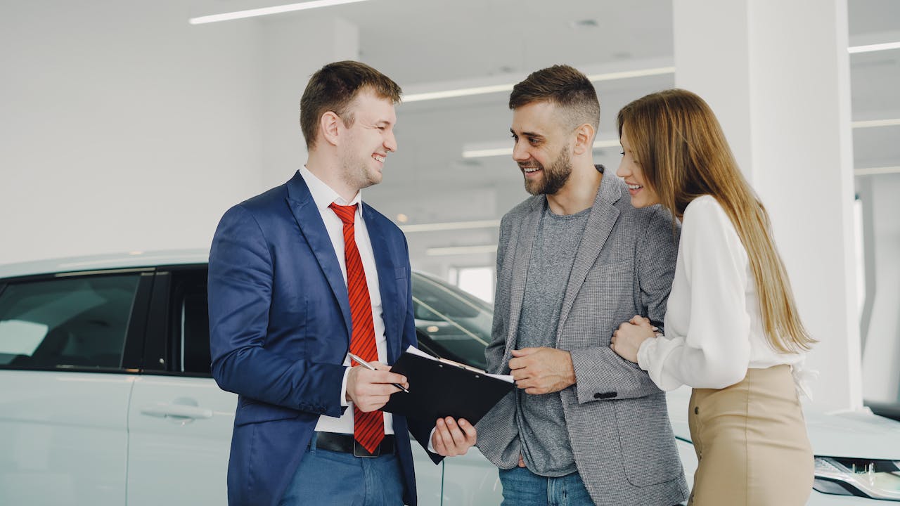 About A smiling couple buys a new car from a confident salesman inside a modern car dealership.