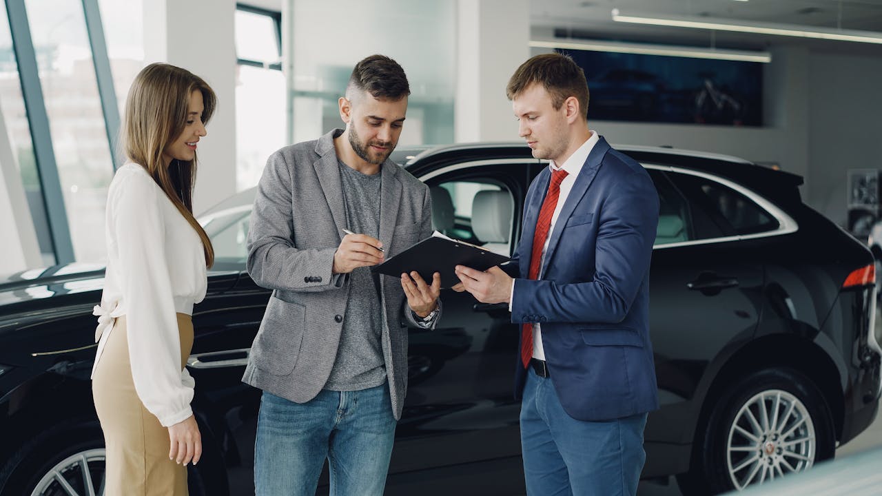Home Three adults discussing documents at a car dealership beside a black car.