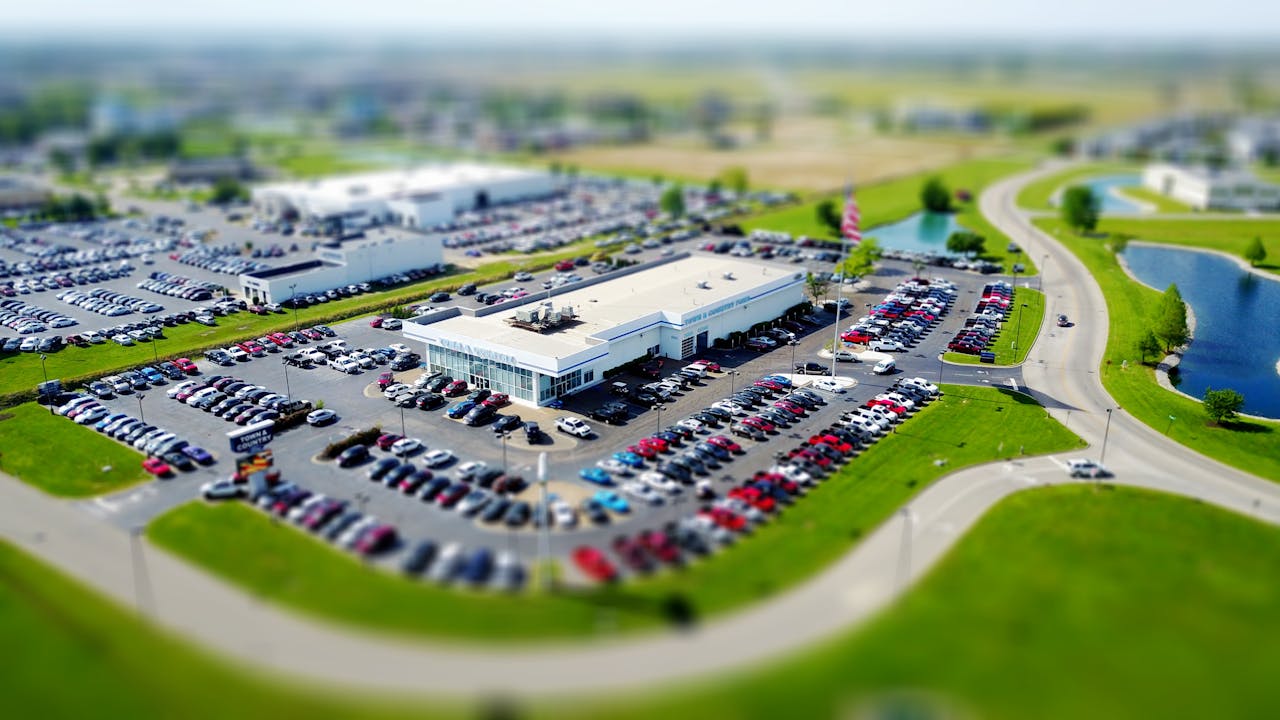 Home Aerial high-angle view of a bustling car dealership surrounded by parked cars in a green landscape.
